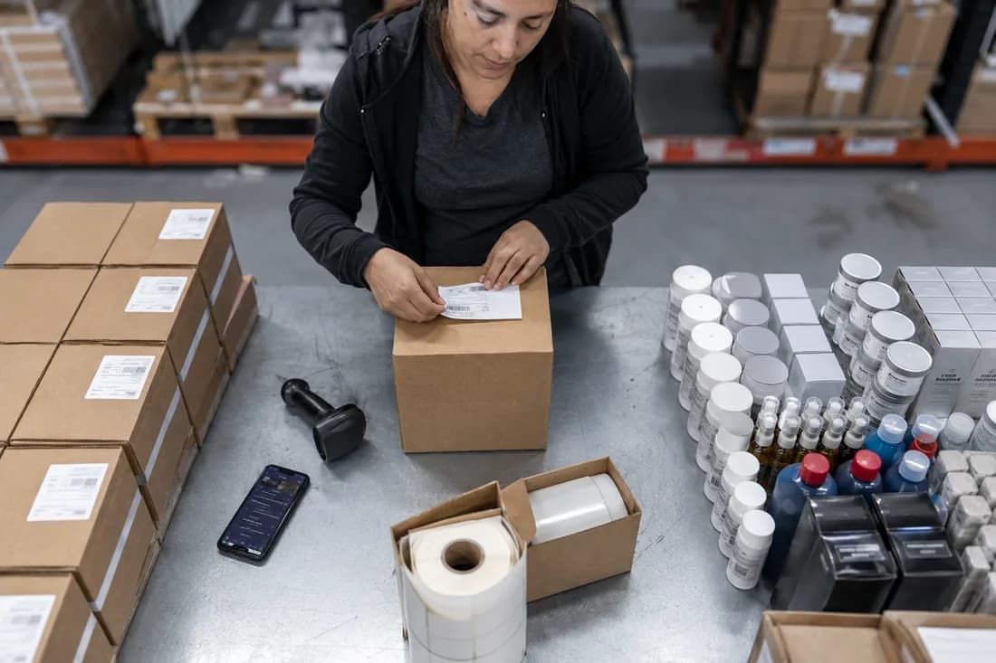 Operaria de un depósito etiquetando una caja por bulto, con el lector de código de barras y el celular sobre la mesa de trabajo